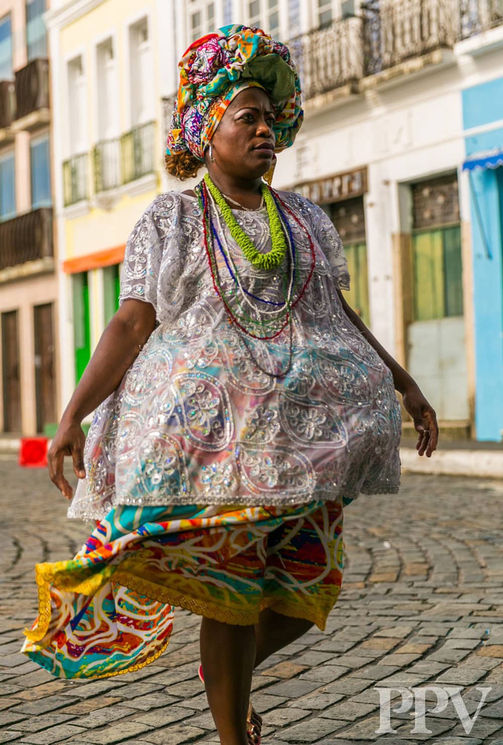 Vestido tradicional colorido de una mujer con tocado y aretes, en una calle de ciudad, mostrando riqueza cultural en la vestimenta y accesorios tradicionales.